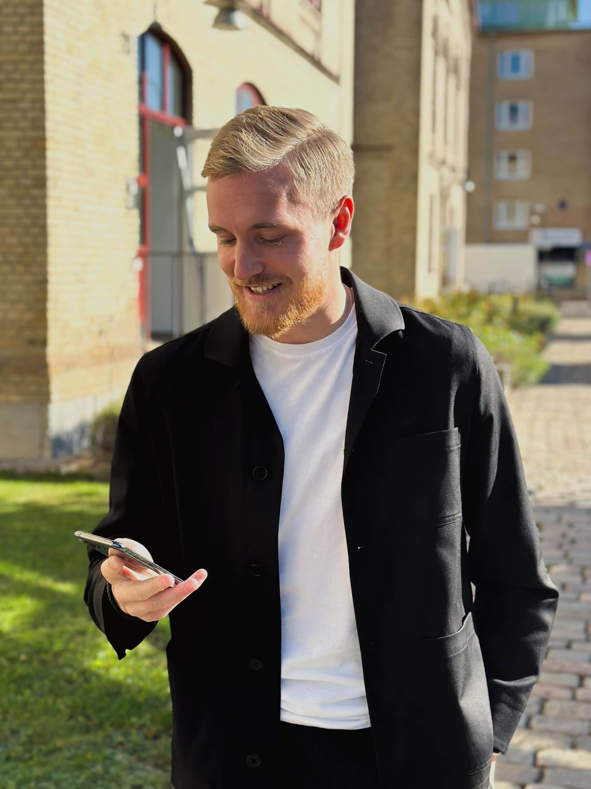 Ung man med mobil i stadsmiljö, ler och tittar på skärmen.