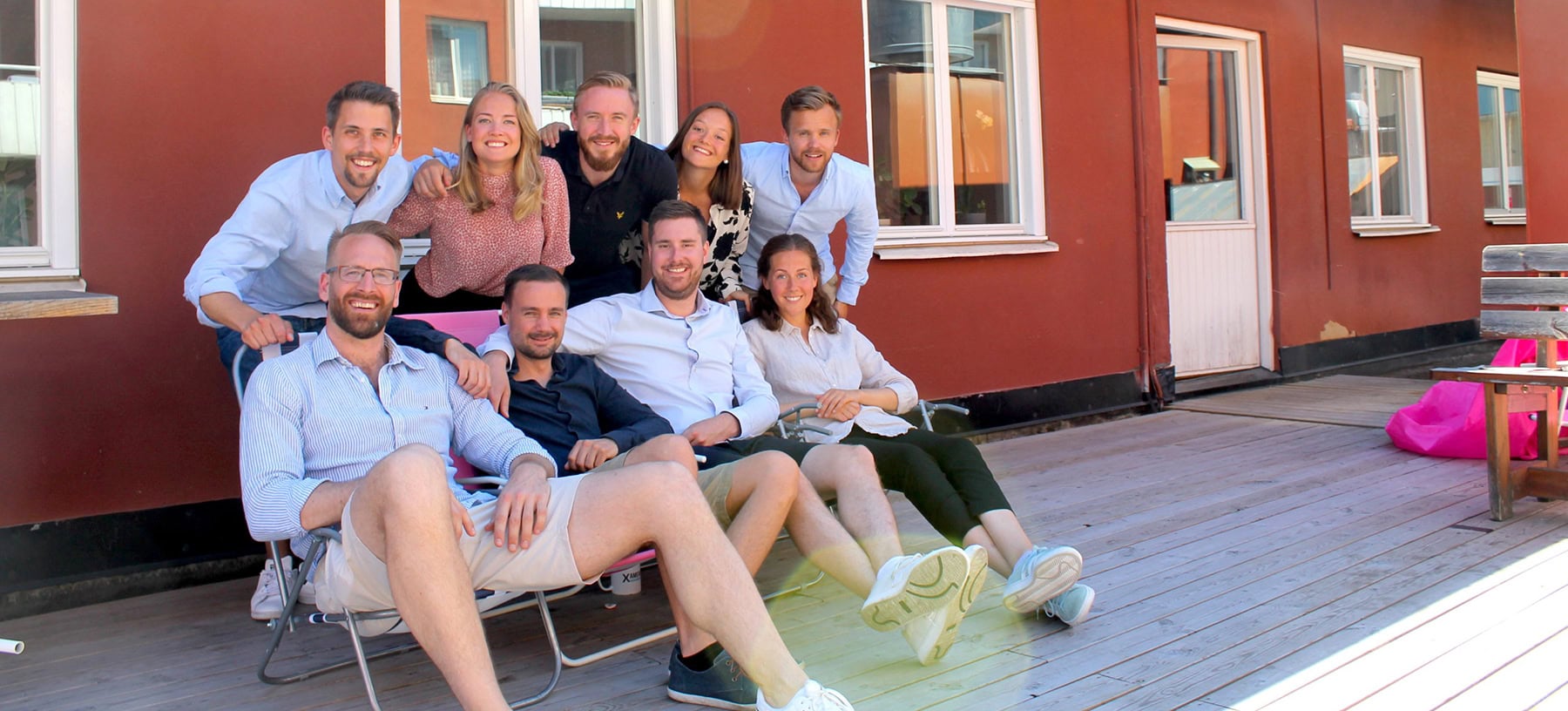 Grupp av unga vuxna som sitter och st&aring;r p&aring; en uteplats utanf&ouml;r en r&ouml;d byggnad.